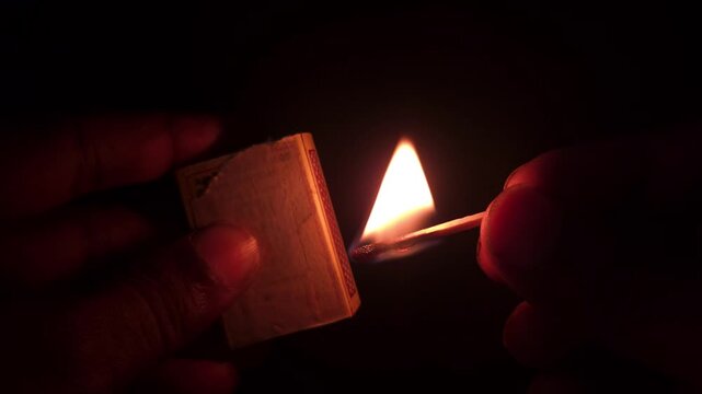 Extreme close-up captures fingers lighting a match, the sulfur tip igniting with fire and smoke, burning brightly in the darkness.