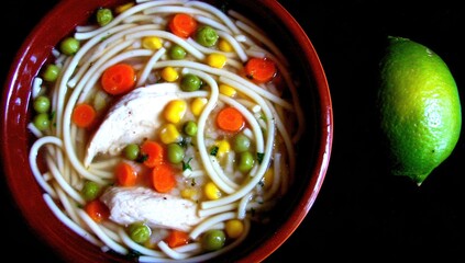 Chicken noodle soup in a red bowl, topped with vegetables and a lime