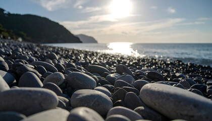 Close Up View Of Smooth Gray Pebbles At A Rocky Beach With Sunlight Sparkling On The Water And Green Hillside In The Background