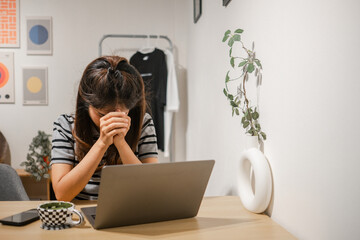 A woman sits at a desk with a laptop, appearing stressed, in a modern home office environment.