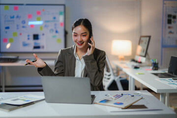 Young manager working late using laptop and talking on smartphone in modern office with whiteboard,...