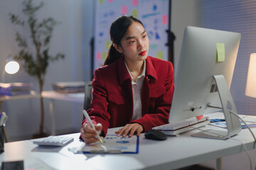 Asian businesswoman working late at night in office analyzing financial charts on computer and...