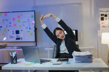 Asian businesswoman stretching arms while sitting at office desk working late at night, overwork and overtime concept