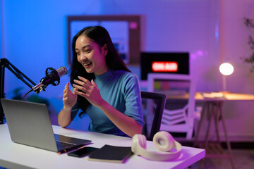 Asian podcaster speaking into microphone recording audio podcast in home studio at night, using professional microphone and laptop