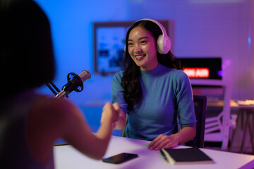 Two female podcasters shaking hands while sitting at table with microphone and headphones in recording studio with neon lights