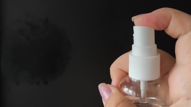 A woman's hand presses the button of a transparent plastic spray bottle and sprays water on a black background. Spray, perfume.