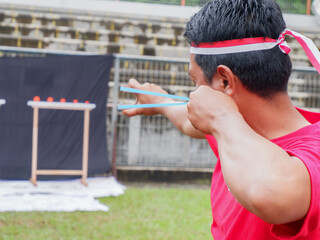 A man wearing a red shirt and Indonesian flag headband is playing a traditional competition during Independence Day celebration, aiming a slingshot at targets outdoors in a festive game.