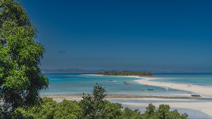Fototapeta premium A sandbar winds through the aquamarine ocean to a small tropical island. Silhouettes of boats, people on the beach. A frame of green trees in the foreground. Clear blue sky. Copy space. Madagascar.