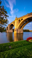 Fototapeta premium Serene sunset view of a stone arch bridge spanning a calm river, reflected in the water, with lush green banks and autumnal foliage