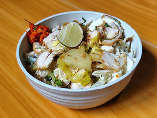 A bowl of Indonesian chicken soto with shredded chicken, sliced boiled egg, vermicelli, sliced cabbage, bean sprouts, celery, chili sauce and lime, mixed with delicious sauce. On a wooden table.	
