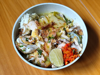 A bowl of Indonesian chicken soto with shredded chicken, sliced boiled egg, vermicelli, sliced cabbage, bean sprouts, celery, chili sauce and lime, mixed with delicious sauce. On a wooden table.	
