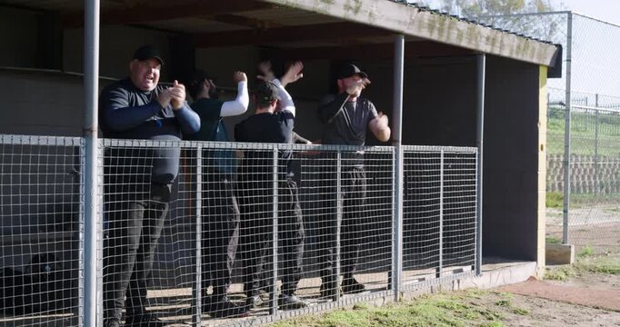 Fist-pumping after play, diverse male coaches behind chain-link railing in dugout cheering on team