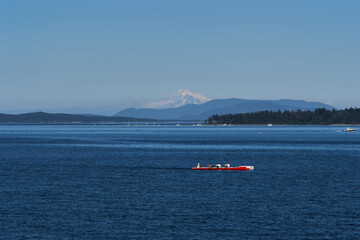 Obraz premium A red rowing boat glides across the ocean near Sidney, Vancouver Island, with distant mountains and Mount Baker in the background.