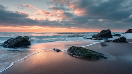 A serene coastal scene captures waves gently lapping against rocks at sunset, with soft orange hues reflecting on wet sand under a partly cloudy sky.