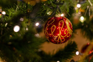 Red and gold ornament hangs from a Christmas tree. The ornament is shaped like a ball and has a gold design on it. The tree is lit up with lights, creating a warm and festive atmosphere
