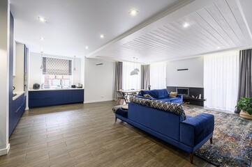 Bright open-plan living space with a blue sofa, kitchen, dining area, and fireplace. Modern design with wooden floors and white ceiling