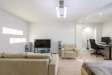 bright, modern living room with a sofa, TV, speakers, armchair, and a desk area. White walls and wooden floor create a clean, minimalist space