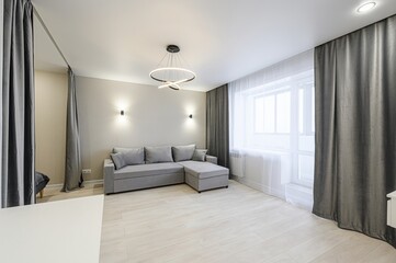 modern, well-lit living space with a gray sofa, curtains, a statement chandelier, and neutral tones. A bedroom area is subtly separated by curtains