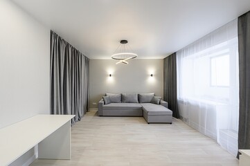 Modern living room with a grey sofa, curtains, and a unique ring chandelier. Minimalist design with light wood floors and neutral walls