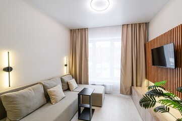 cozy living room features a beige sofa, a TV on a wood slat wall, neutral curtains, and modern lighting. The room is bright and appears stylish