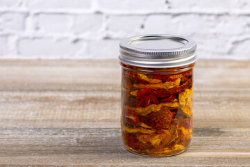 A glass jar with sun dried red and yellow cherry tomatoes in olive oil and with herbs. Jar on the rustic brown wooden table.