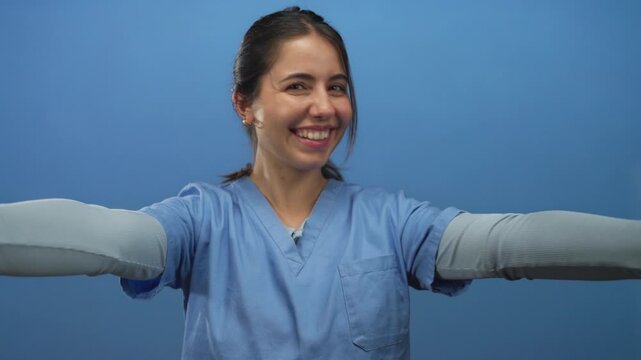 Woman smiling in hospital scrubs against isolated blue background showing arms outward in a welcoming gesture representing healthcare positivity and confidence.