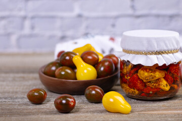 Glass jar with dried red, black and yellow cherry tomatoes in olive oil and with herbs on the rustic brown wooden table. Various cherry tomatoes in a wooden plate and white towel on the background.