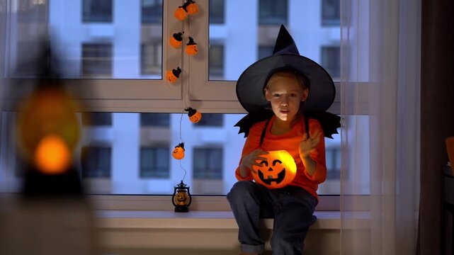 Happy girl sits by window in witch hat and conjures over halloween pumpkin lantern in evening. Child celebrates halloween