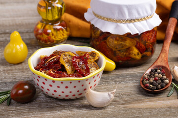 Jar with dried red and yellow tomatoes in olive oil and with herbs on the rustic brown wooden table. Various cherry tomatoes, spoon with peppercorns, rosemary, garlic and yellow towel around it.