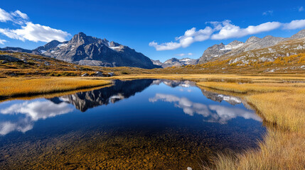 Majestic mountains reflect on calm lake surrounded by golden grasslands and blue skies