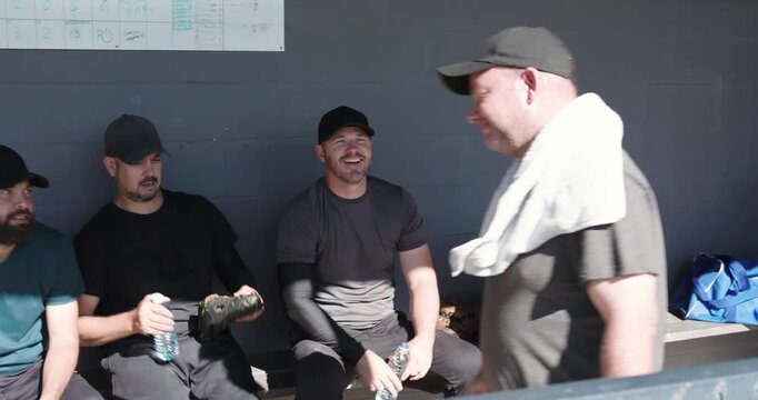 Second player passing bottle motivating Diverse male teammates hydrating in dugout under scoreboard