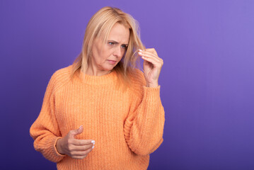 Concerned middle-aged woman in orange sweater looking at her hair with frustration. Concept of stress, aging, hair loss, hormonal issues, and emotional expression in daily life.