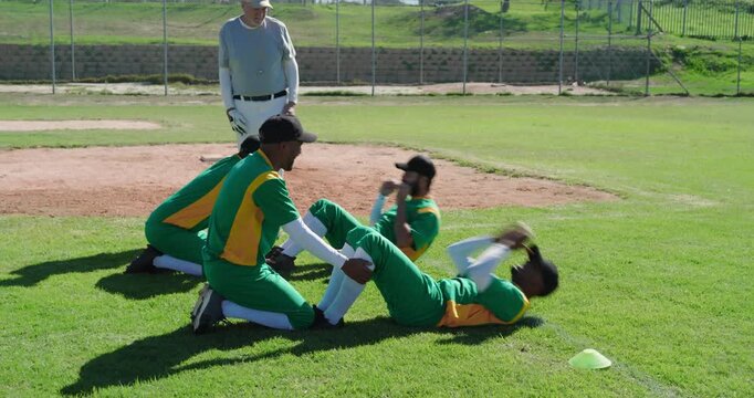 Entering male coach instructing teammates holding South Asian man doing sit-ups infield, with glove