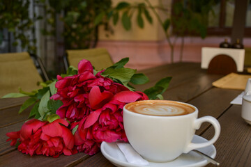 Romantic morning coffee with latte art and fresh red peonies on wooden table