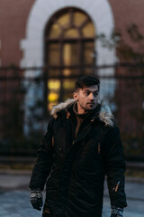 Fototapeta premium Man in black winter coat with fur hood and gloves standing outdoors near brick building. Warm clothing, urban autumn fashion, casual walk, modern city style, architectural background