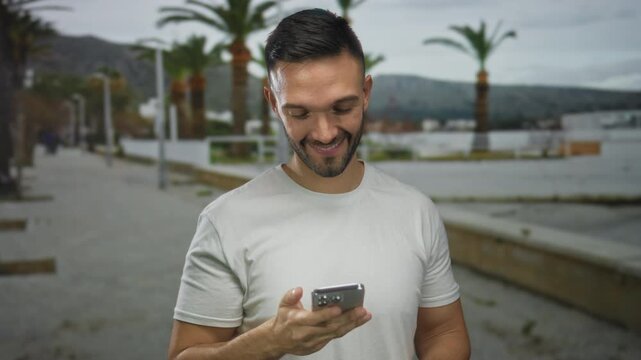 Young man smiling at phone while walking on promenade near beach with palm trees in background on a cloudy day outdoors.