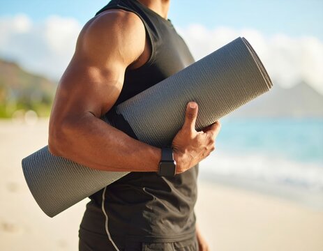 A muscular man holding a yoga mat on the beach, ready for a workout.