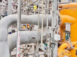 An intricate network of industrial pipes and machinery in a factory setting. A large yellow tank is visible, along with multiple valves, gauges, and electrical components.