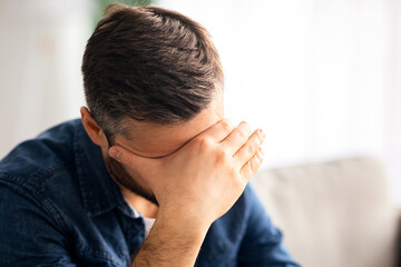 Upset middle-aged bearded man having headache, sitting on couch at home and touching his forehead, copy space. Closeup of frustrated man suffering from difficulties or depression, copy space