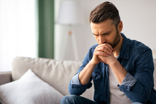 Upset bearded middle-aged man in casual outfit sitting on sofa in living room, thinking about something, looking down, frustrated man having financial problems during pandemic, copy space