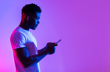 Side view of young black man in earphones listening to music, using new mobile app on smartphone in neon light, free space. Cheerful African American guy enjoying new playlist or audio book