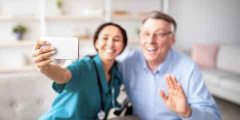 Cheerful disabled senior man in wheelchair and his nurse taking selfie indoors, selective focus. Impaired elderly patient with young doctor making mobile photo of themselves at retirement home