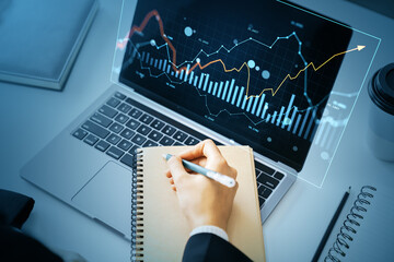 Businessperson analyzing financial chart data on laptop with digital graph interface and taking notes in notebook on office desk.