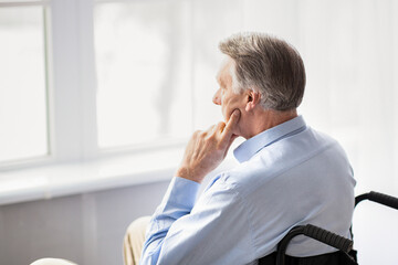 Fototapeta premium Depression in disabled seniors. Upset elderly man in wheelchair shutting himself off from world, looking out window at home. Handicapped older patient feeling unhappy and lonely indoors