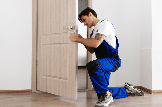 Professional Repair Service Concept. Focused young male locksmith workman in blue uniform installing door knob. Installation of a lock on the front wooden entrance door at home or office workplace