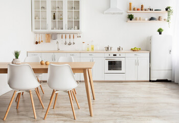 Light kitchen design in scandinavian style. Wooden dining table and white furniture with different utensils and accessoires on shelves, small white refrigerator near window, empty space