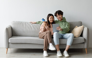Millennial Asian couple with credit card and digital tablet sitting on sofa, ordering goods on internet, buying things in web store at home, copy space. Online shopping, remote banking concept
