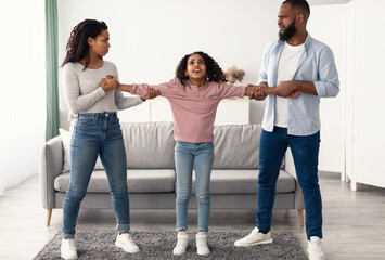 Divorce Concept. Portrait of angry African American parents fighting over their child, mad man and woman quarrelling, pulling daughter's hands in different directions. Domestic Violence, Separation
