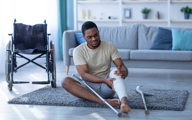 Young black man with injured leg sitting on floor after falling down, having trouble walking with...