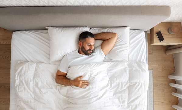 Top view of sleeping middle aged guy enjoying peaceful restful nap in his bed, holding hand behind head, in bedroom interior. Sleep and relaxation for health concept - Powered by Adobe